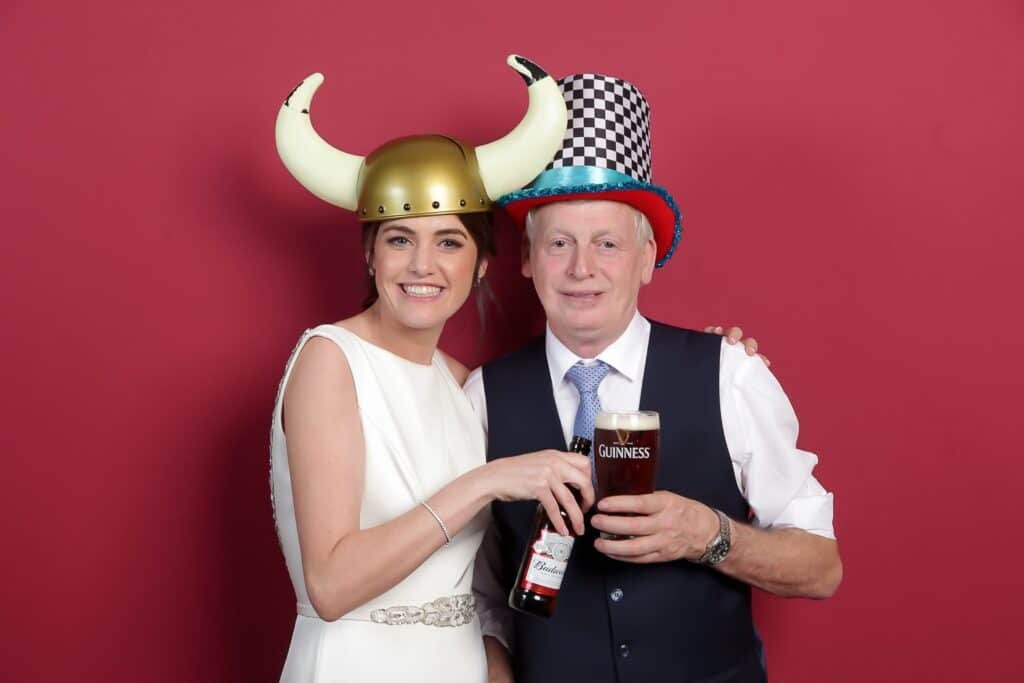 A woman in a white dress and a horned helmet smiles while holding a bottle. A man in a checkered hat and vest holds a pint of Guinness. Both stand against a red background.