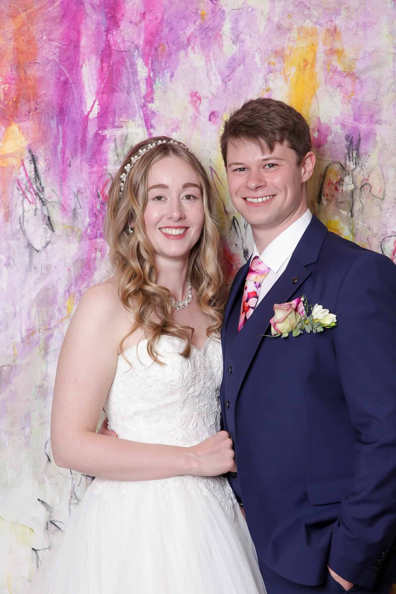 A bride and groom stand smiling in front of a colorful, abstract background. The bride wears a white dress and pearl headband, while the groom dons a navy suit with a patterned tie and boutonniere.