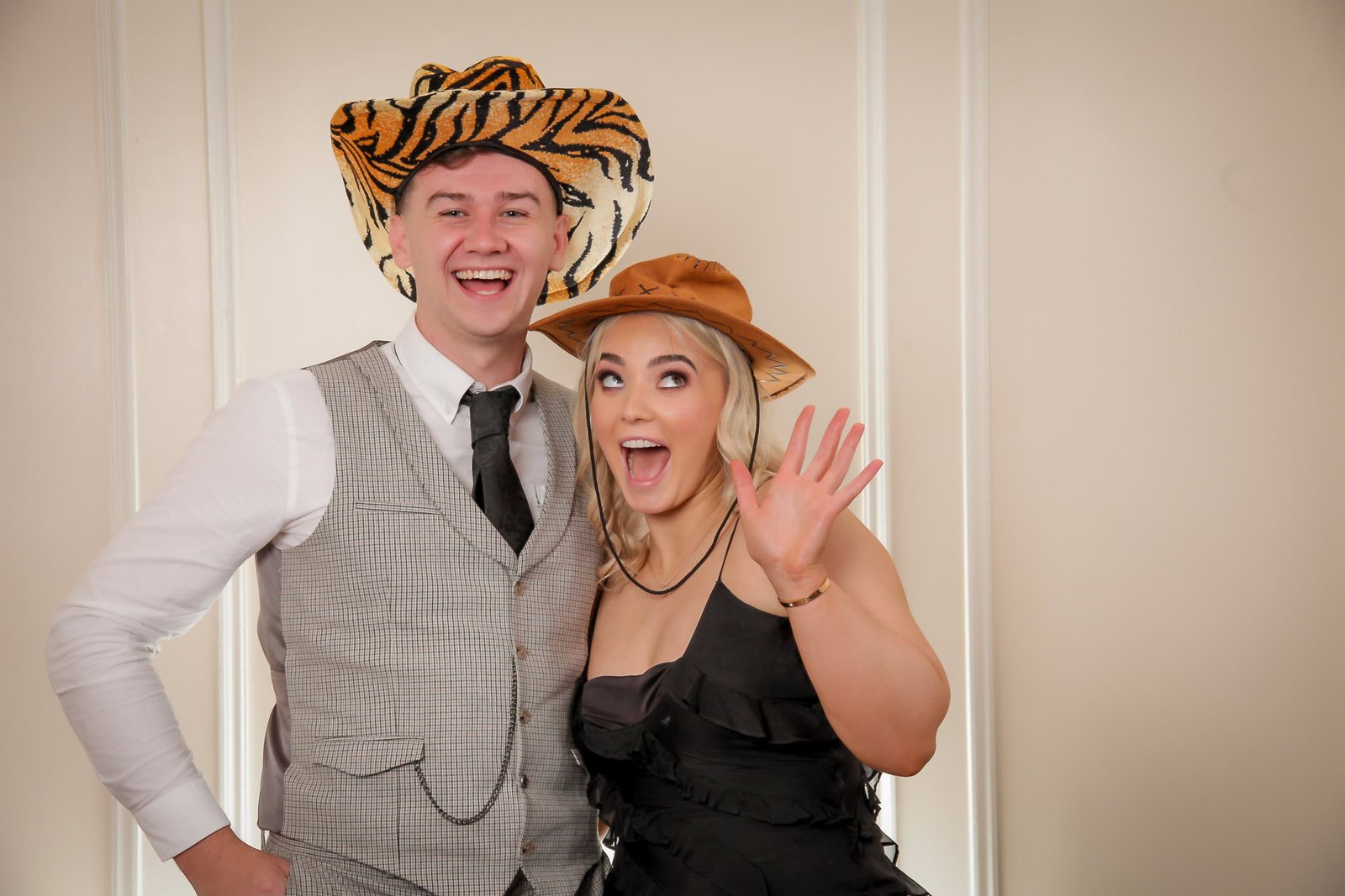 A man and woman stand in front of a beige wall, both smiling. The man wears a gray vest, white shirt, black tie, and a tiger-striped cowboy hat. The woman wears a black dress and a brown cowboy hat, with one hand raised in a wave.