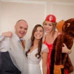 Three people pose indoors. The man on the left wears a vest and holds a veil, the woman in the center wears a white dress, and the woman on the right wears a red helmet and holds a large stuffed monkey. They are smiling and standing close together.