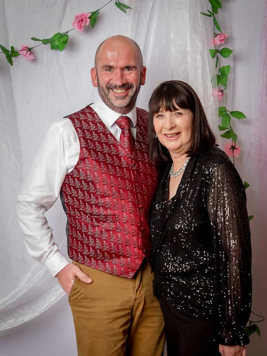 A man and a woman standing together, smiling. The man is wearing a red patterned vest, white shirt, and brown pants. The woman is in a black sequined top and black pants. They are in front of a white draped background with pink roses and green leaves.