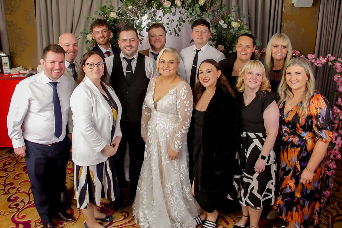 A group of people, including a bride in a white dress and a groom in a dark suit, pose for a photo at a wedding. They are standing on a patterned carpet with floral decorations in the background. Everyone is dressed in formal attire.