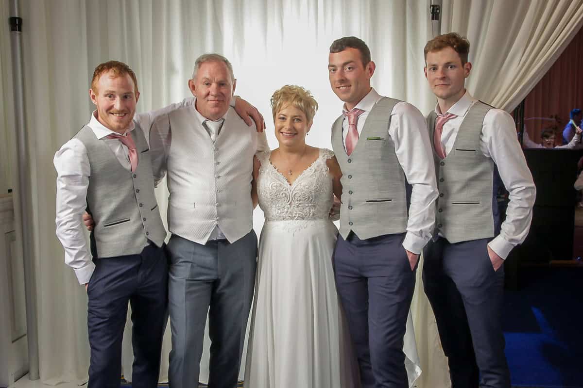 A wedding party poses together in front of a white curtain. The group consists of four men and one woman in the center, wearing wedding attire. The men wear gray vests and pink ties, and the woman wears a white gown.