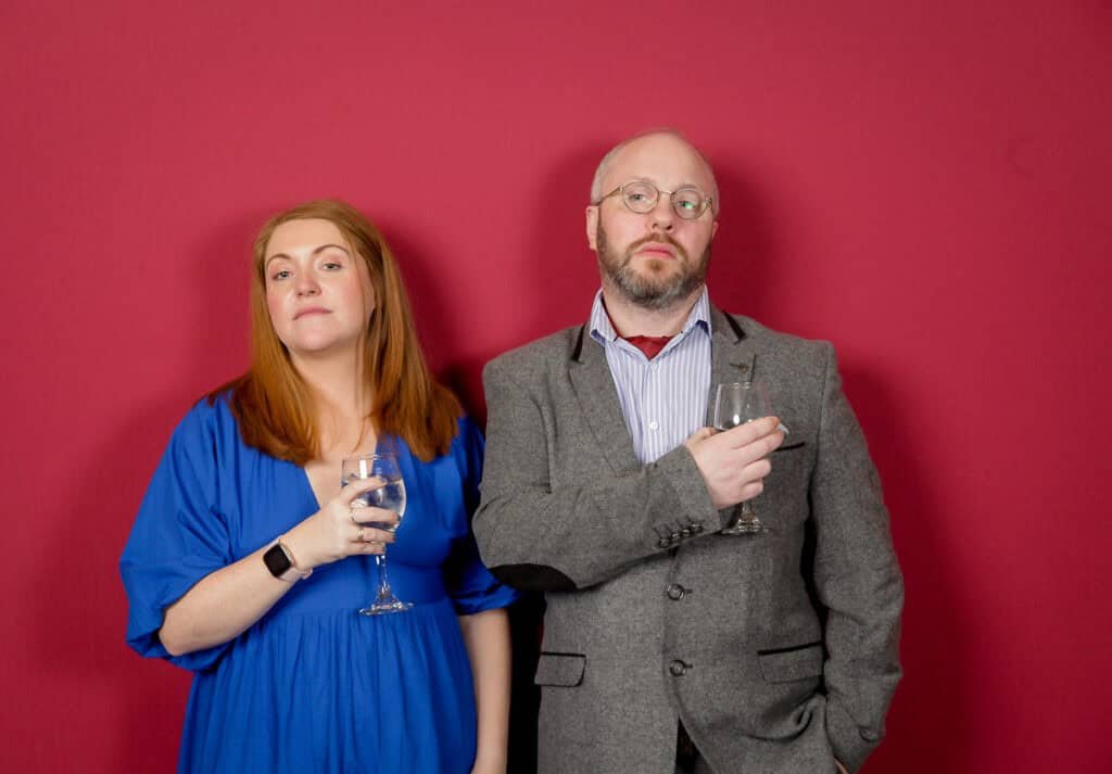 A man and woman stand against a red background, each holding a wine glass. The woman has long red hair and wears a blue dress. The man has short hair, a beard, and wears glasses, a gray suit, and a striped shirt. Both have a relaxed expression.