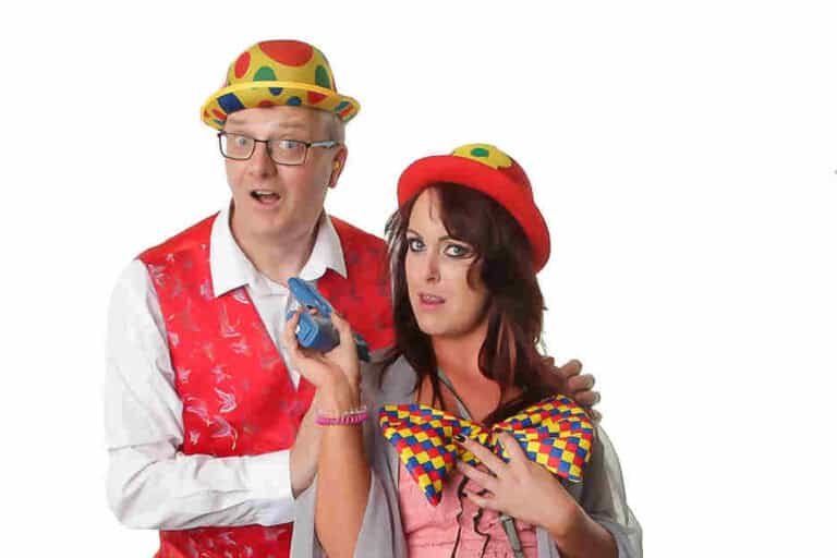 Two people wearing colorful clown costumes pose against a white background. The man wears a hat and a red patterned vest, and the woman holds a blue object and wears a bright bow tie and hat.