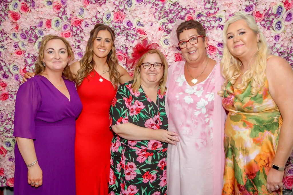Five women stand in front of a floral backdrop. They are wearing colorful dresses in shades of purple, red, and floral patterns. One woman wears a red fascinator. They are smiling and posing for the photo.