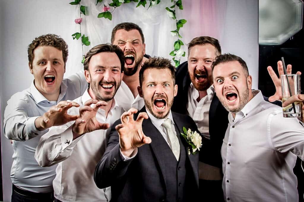 Six men in dress shirts and suits are posing together in a playful, roaring gesture. The man in the center is wearing a suit with a white boutonniere. A floral backdrop is behind them, suggesting a celebratory event.