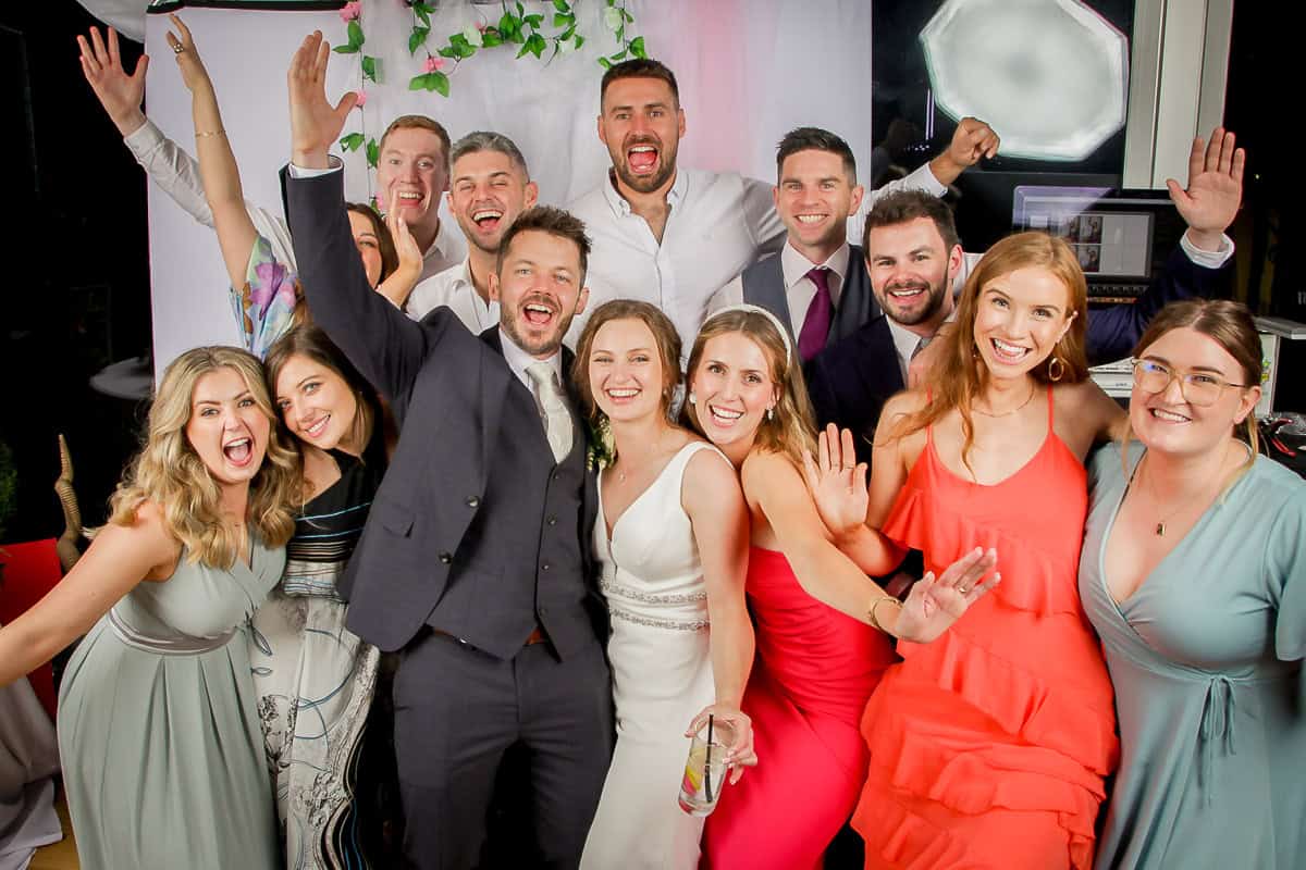 A group of people, dressed in formal attire, smiling and posing for the camera. Some are raising their hands in celebration. There is a white backdrop decorated with flowers behind them.