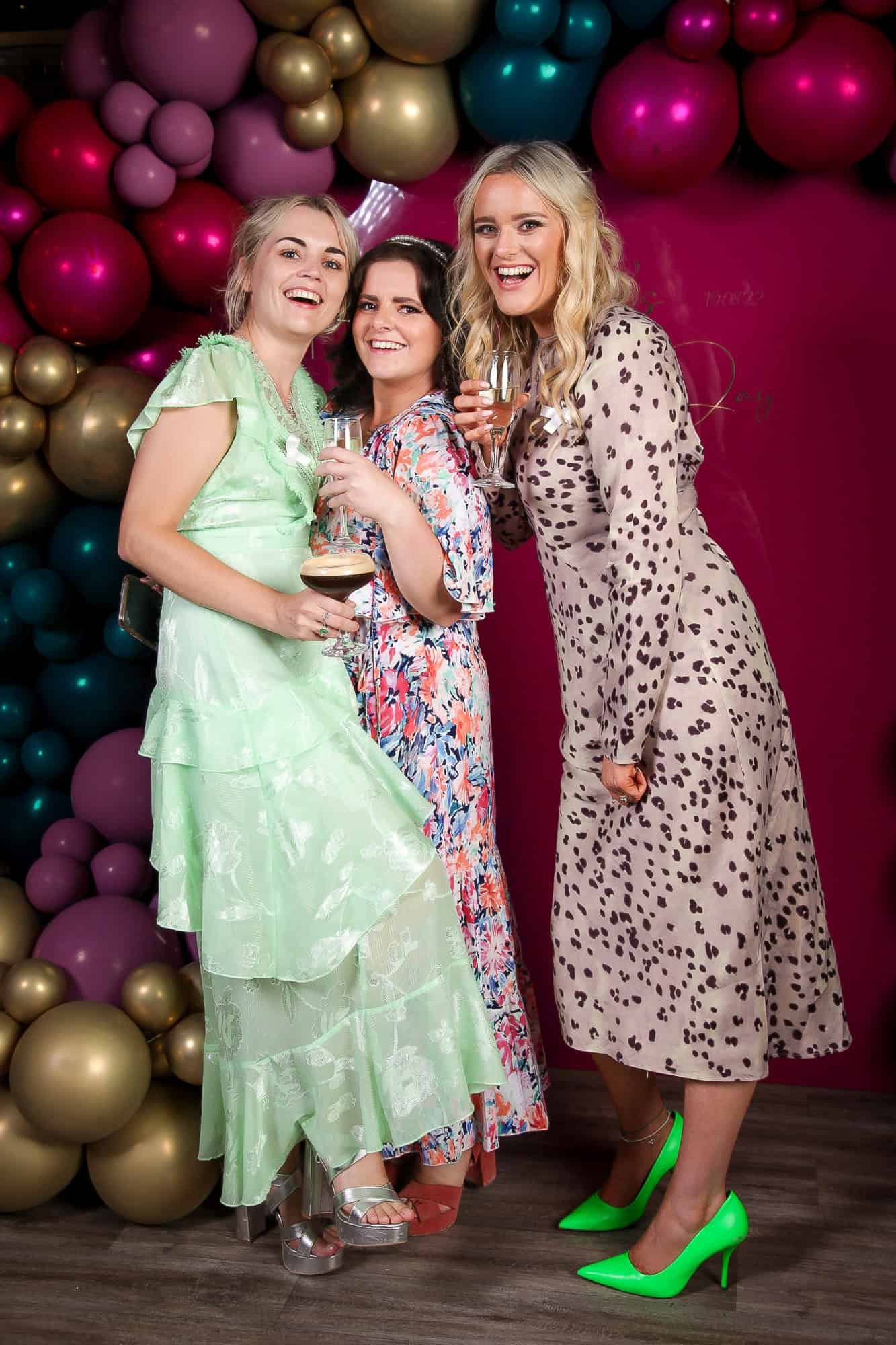 Three women are smiling and holding drinks. They are standing in front of a colorful balloon arrangement featuring gold, pink, and teal balloons. One woman wears a green dress, another a floral dress, and the third a leopard print dress with bright green heels.