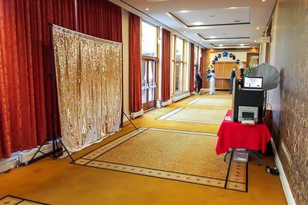 A long hallway with gold carpet and red curtains featuring a photo booth setup. There is a backdrop on the left and a small table with equipment on the right. In the background, a balloon arch is visible near a windowed exit.