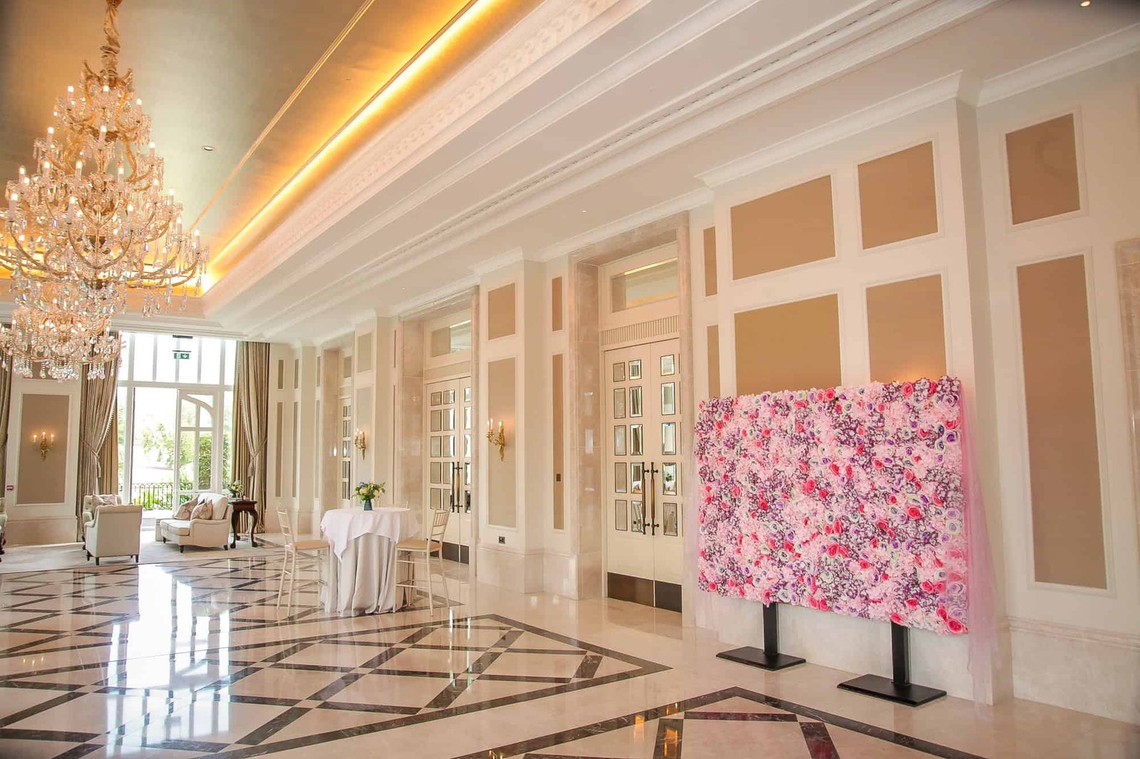 A spacious hotel lobby features a large chandelier, a tiled floor in white and gray patterns, several white armchairs, and a table with a white cloth. A wall displays a large colorful artwork in pink and white hues.