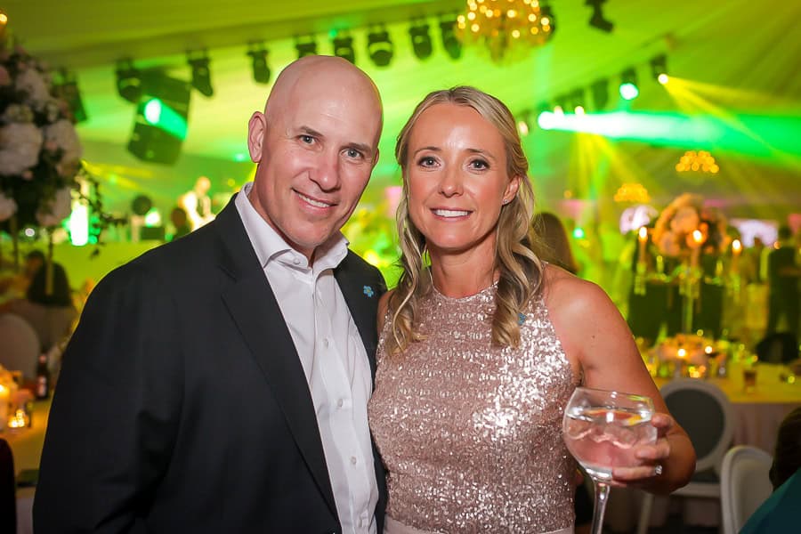 A man in a suit and a woman in a sequined dress stand together at an indoor event. The woman holds a glass, and there are people and green lights in the background.