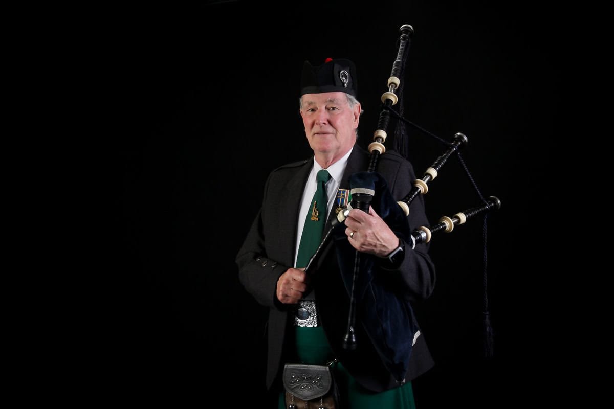 An older man stands against a dark background wearing traditional Scottish attire, including a black jacket, green tie, and kilt. He holds a set of bagpipes at his side.