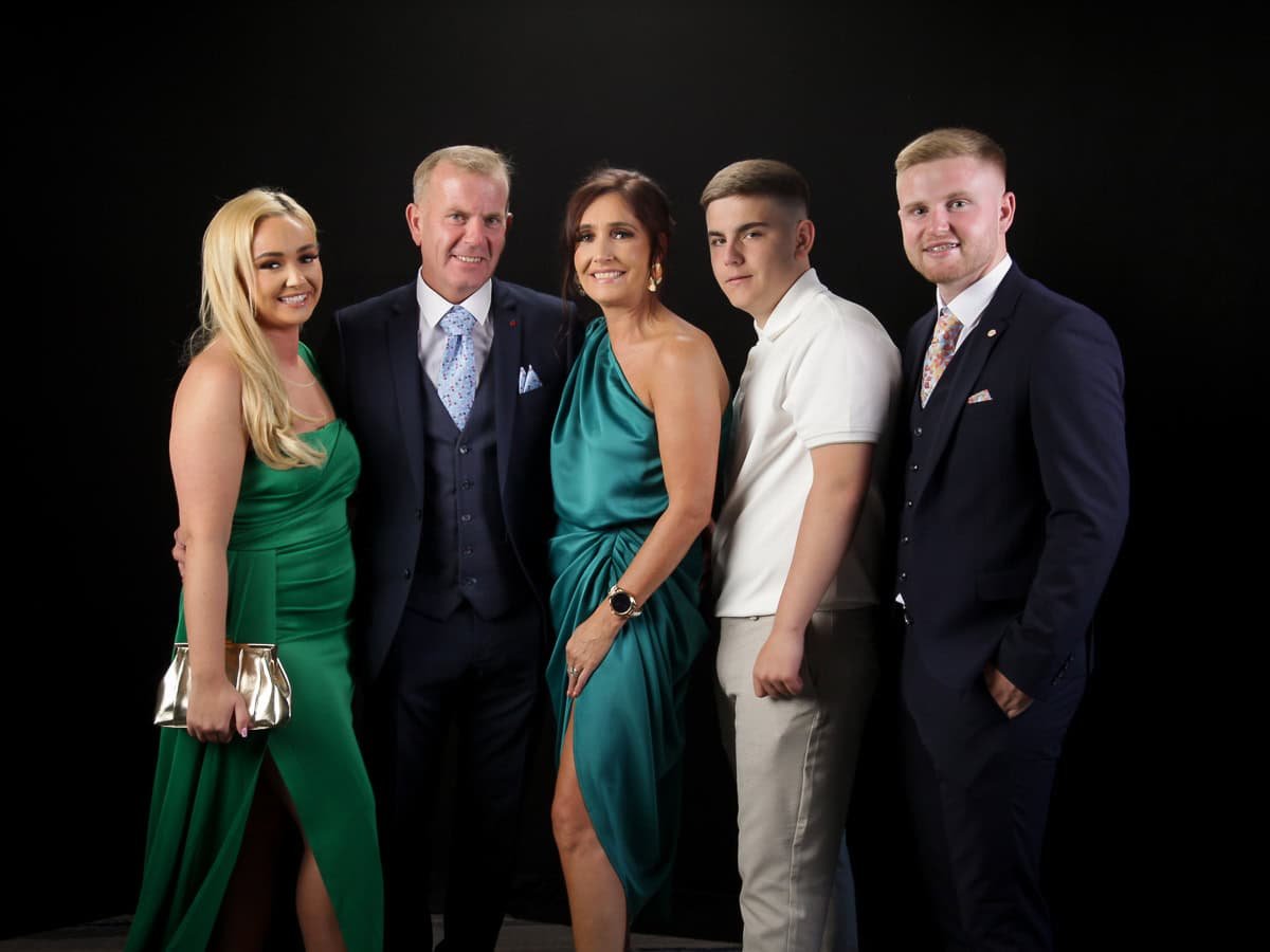 A group portrait with five people dressed in formal attire posing against a dark background. Two women are in green dresses, while three men wear suits, one in a white shirt. They are all smiling at the camera.