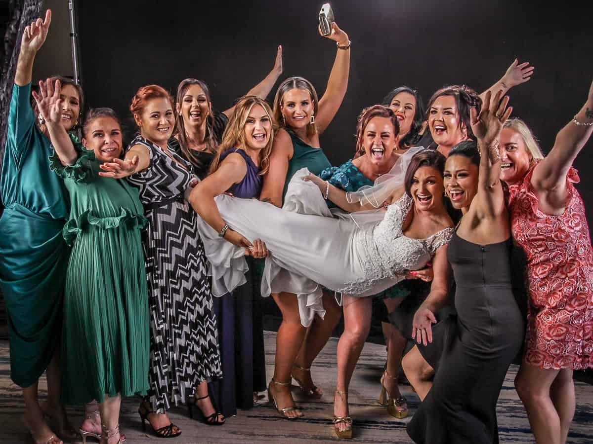 A group of women dressed in various styles and colors of dresses are cheerfully posing together. They are smiling and holding up a woman in a white dress at the center. The background is plain and dark.