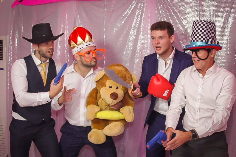 Four men in a playful setting are posing with various props, including a stuffed bear, toy guns, and oversized novelty glasses. They are wearing hats and white shirts, set against a backdrop of shimmering pink fabric.