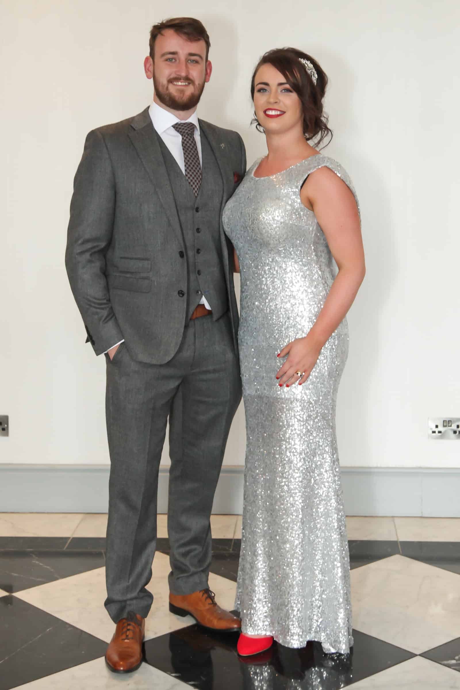 A man in a gray suit and a woman in a silver sequin dress stand on a black and white tiled floor. They are posing for a photo against a plain white wall. The woman is wearing red shoes.