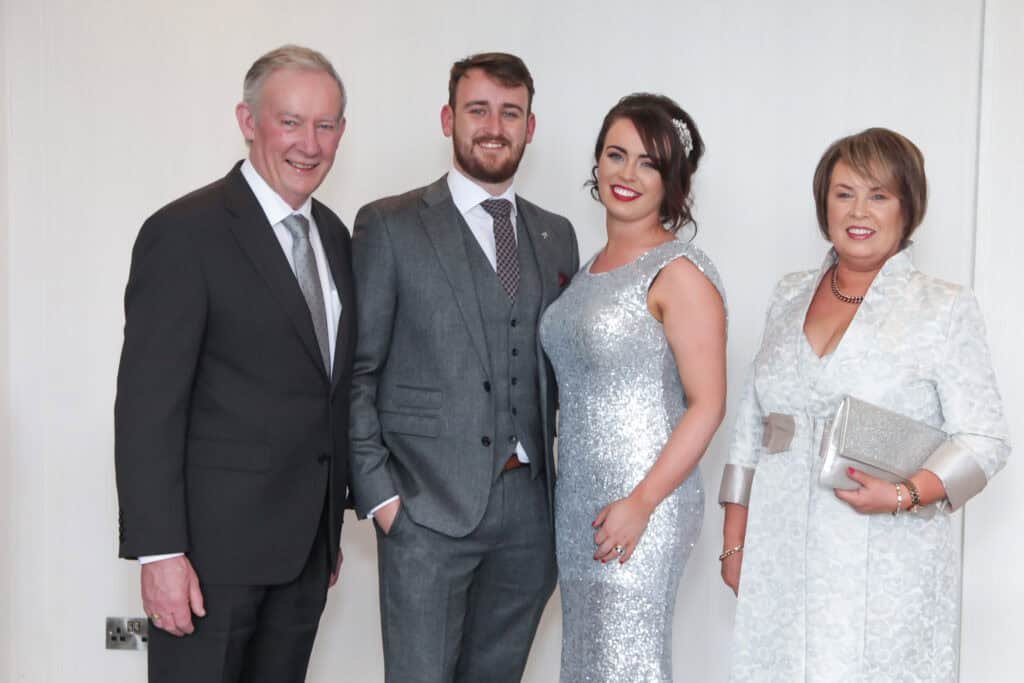 Four people standing indoors dressed in formal attire. The two men are in suits, and the two women are wearing elegant dresses. The woman second from the right is in a silver sequined dress, while the woman on the right wears a light patterned dress.