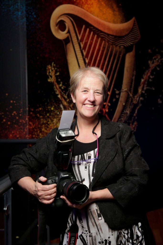 A person with short hair is smiling and holding a camera with a flash attachment. They are wearing a dark jacket over a patterned top. Behind them is a large image of a harp with colorful splashes.