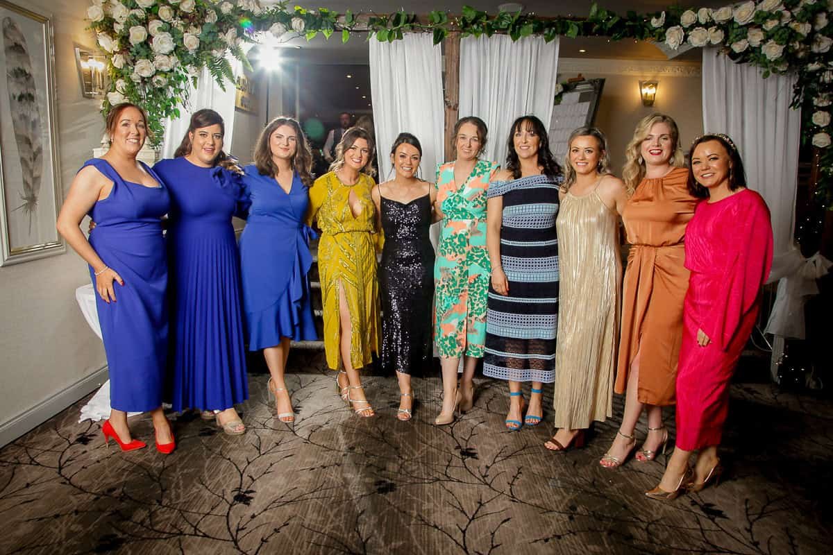 A group of eleven people pose together, standing in a row. They are wearing brightly colored formal dresses. The background features draped white curtains and floral decorations.