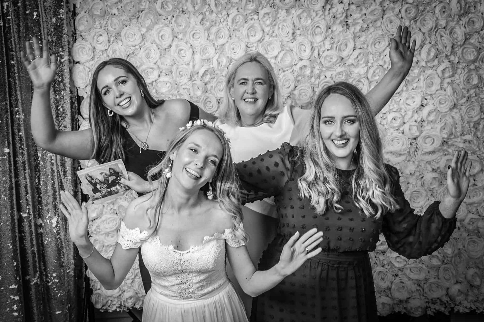 Four women pose joyfully in front of a floral backdrop. The woman on the left holds a small photo. The woman in front is wearing a wedding dress. All appear to be smiling and have their arms raised, expressing excitement. The image is in black and white.