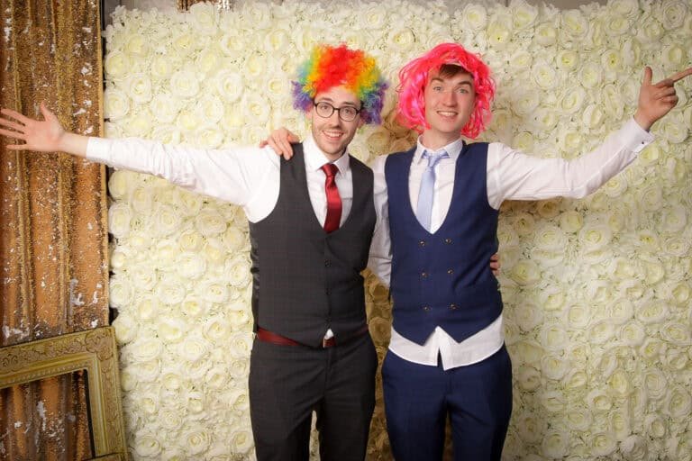 Two people wearing suits stand in front of a white rose backdrop. One wears a rainbow clown wig, and the other wears a pink wig. They both have their arms outstretched and are smiling.