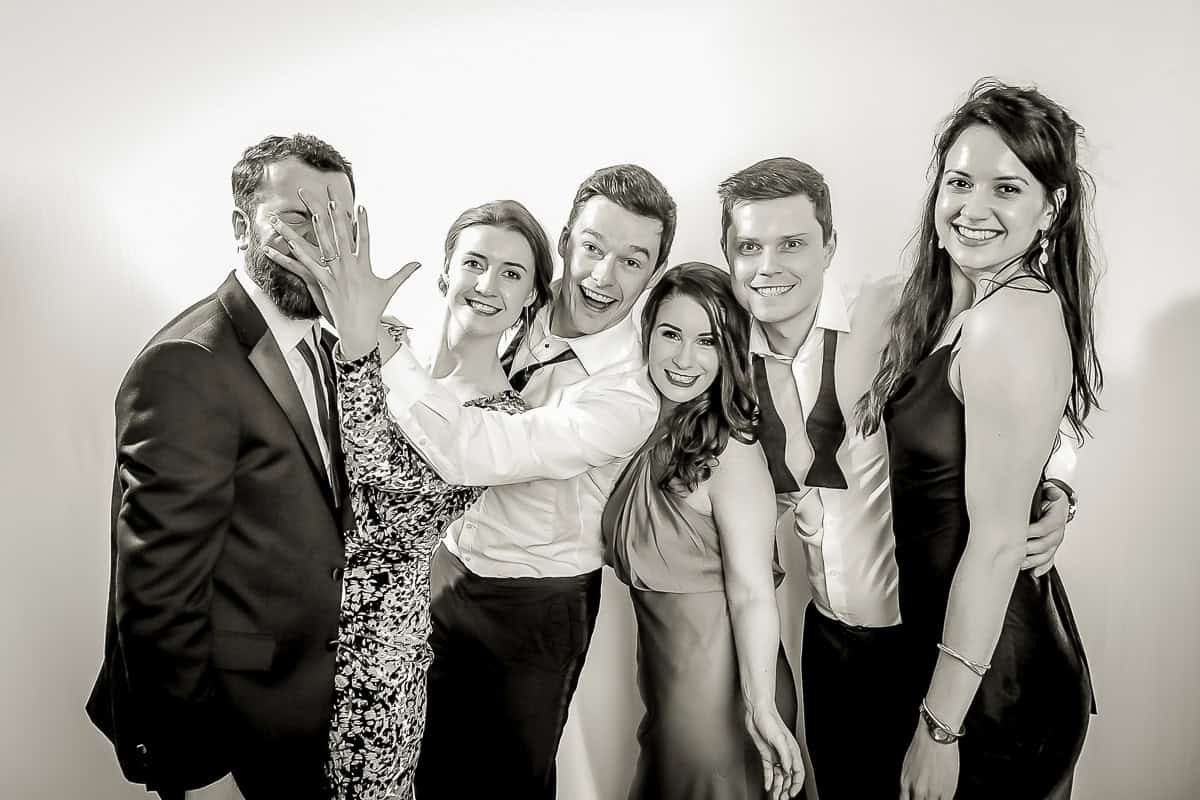 A group of six people pose together, smiling in a playful manner. One person covers another's face with their hand. The group is dressed formally, some in suits and others in dresses, against a plain background. The image is in black and white.