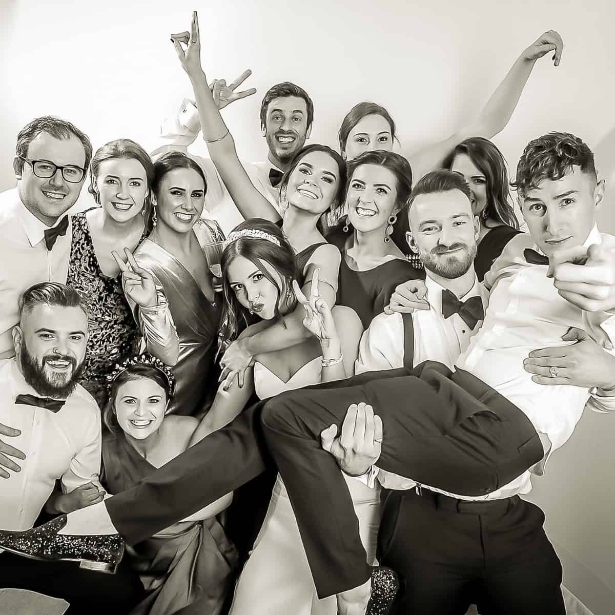 A group of thirteen people posing and smiling at the camera, with playful hand gestures. One person is being held up horizontally by others. They are dressed in formal attire. The image is in black and white.