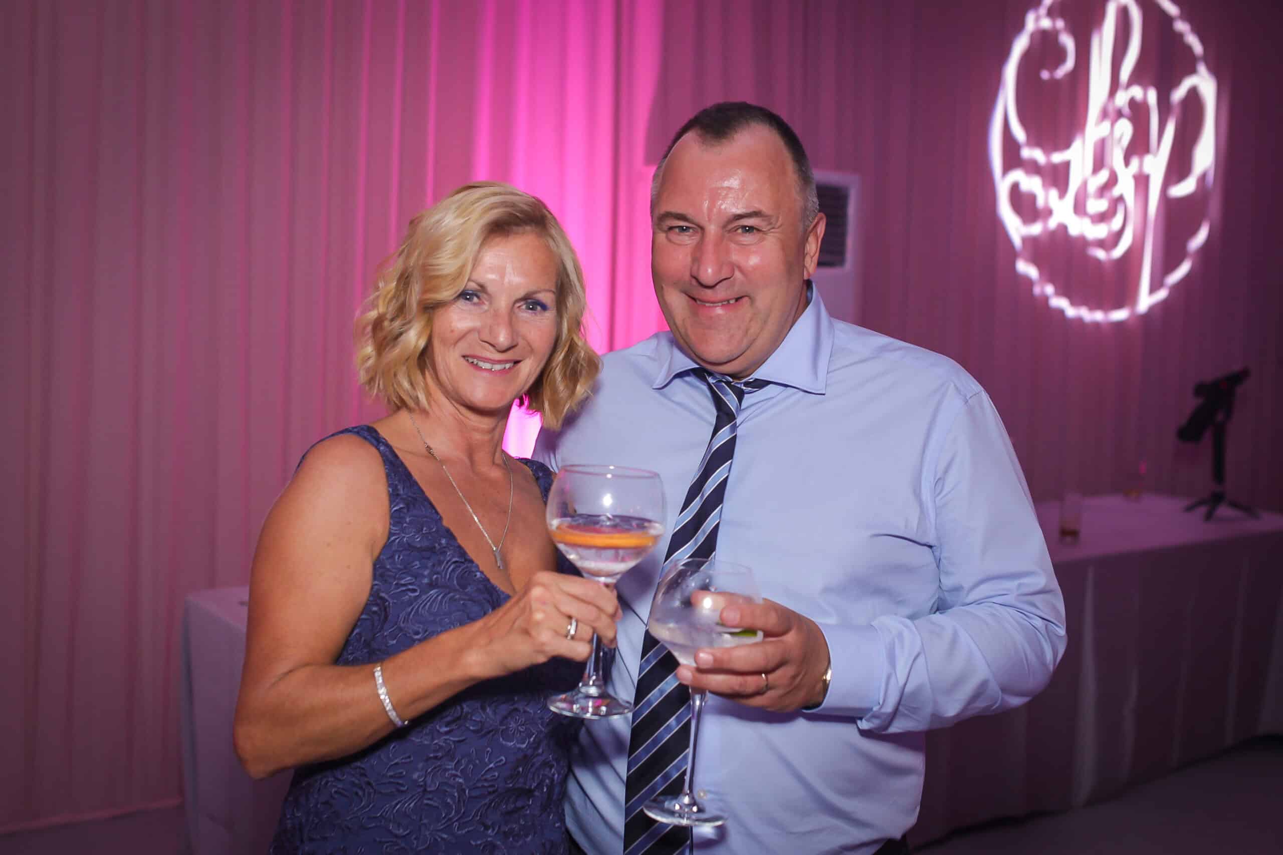A woman in a blue dress and a man in a light blue shirt with a striped tie hold drinks and smile at the camera. They are standing in a room with pink lighting and a monogram decoration on the wall.