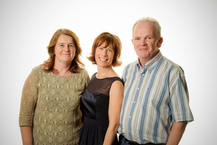 Three adults stand close together against a plain white background. The two women on the left have shoulder-length hair; one wears a lace top, and the other a dark dress. The man on the right wears a striped short-sleeve shirt. They are all smiling.