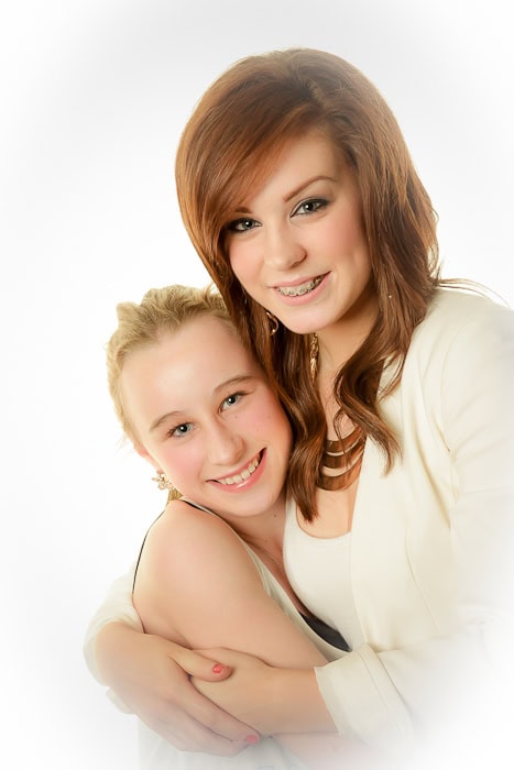 Two smiling females are posing together against a light background. The individual on the right has long auburn hair and braces, while the one on the left has blonde hair pulled back. They are embracing each other closely.