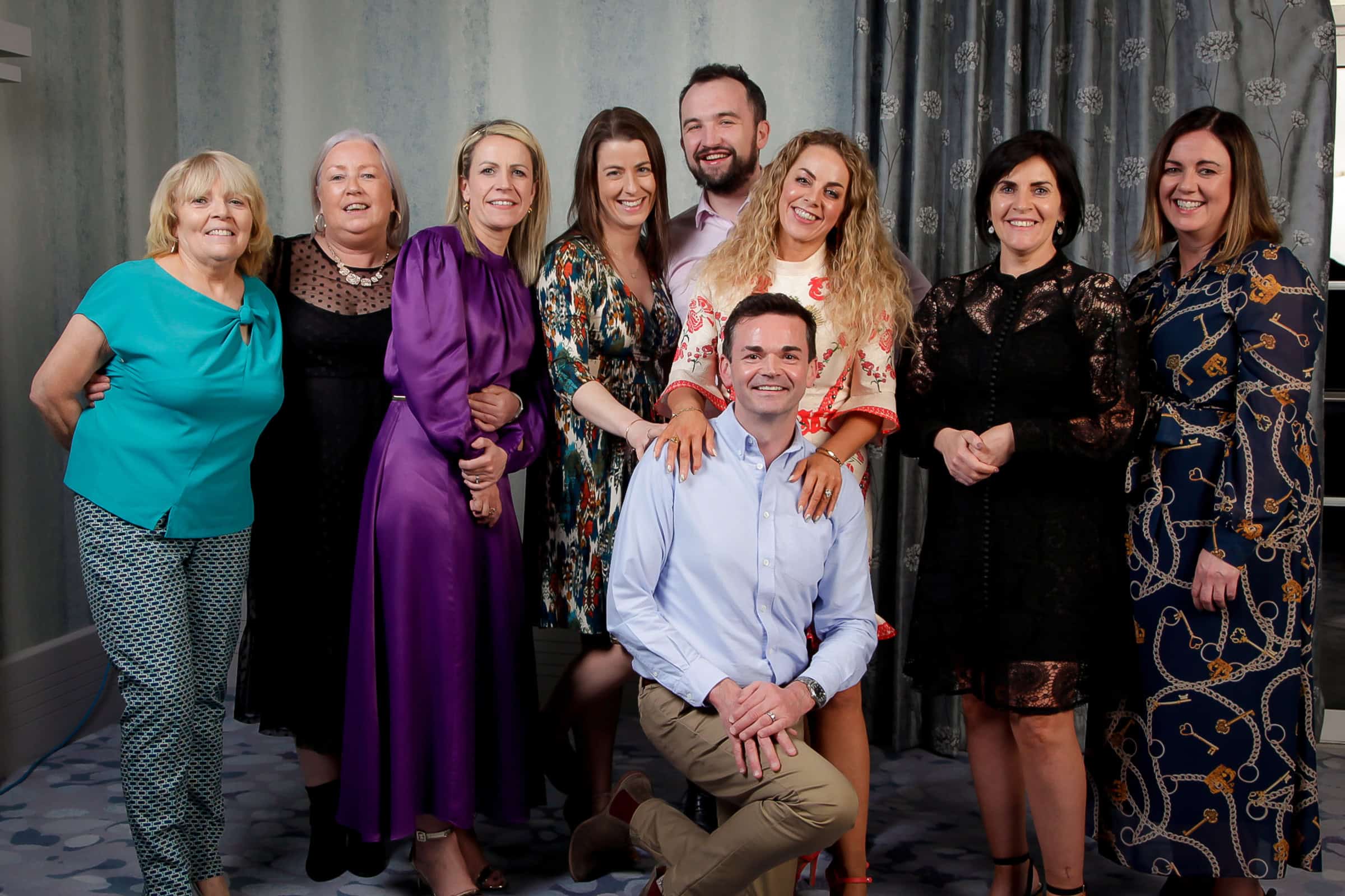 A group of nine people pose for a photograph indoors. Eight are standing while one kneels in front. They are dressed in a variety of outfits, including formal dresses and casual shirts. The background features patterned curtains and a textured wall.