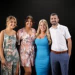 A group of four people stand together against a black background. The group includes three women and one man, all smiling. The women are dressed in colorful dresses, and the man is wearing a white shirt and blue pants.