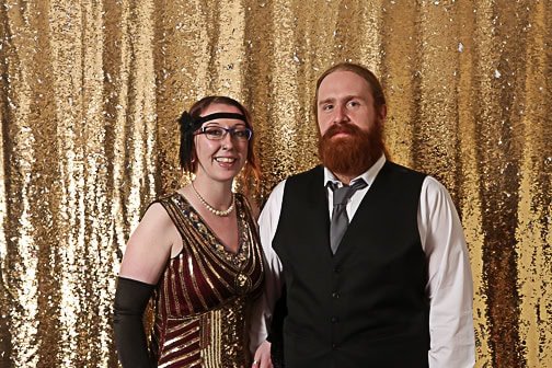 Photo of a smiling couple dressed in vintage attire at a photo booth with a gold sequin background.