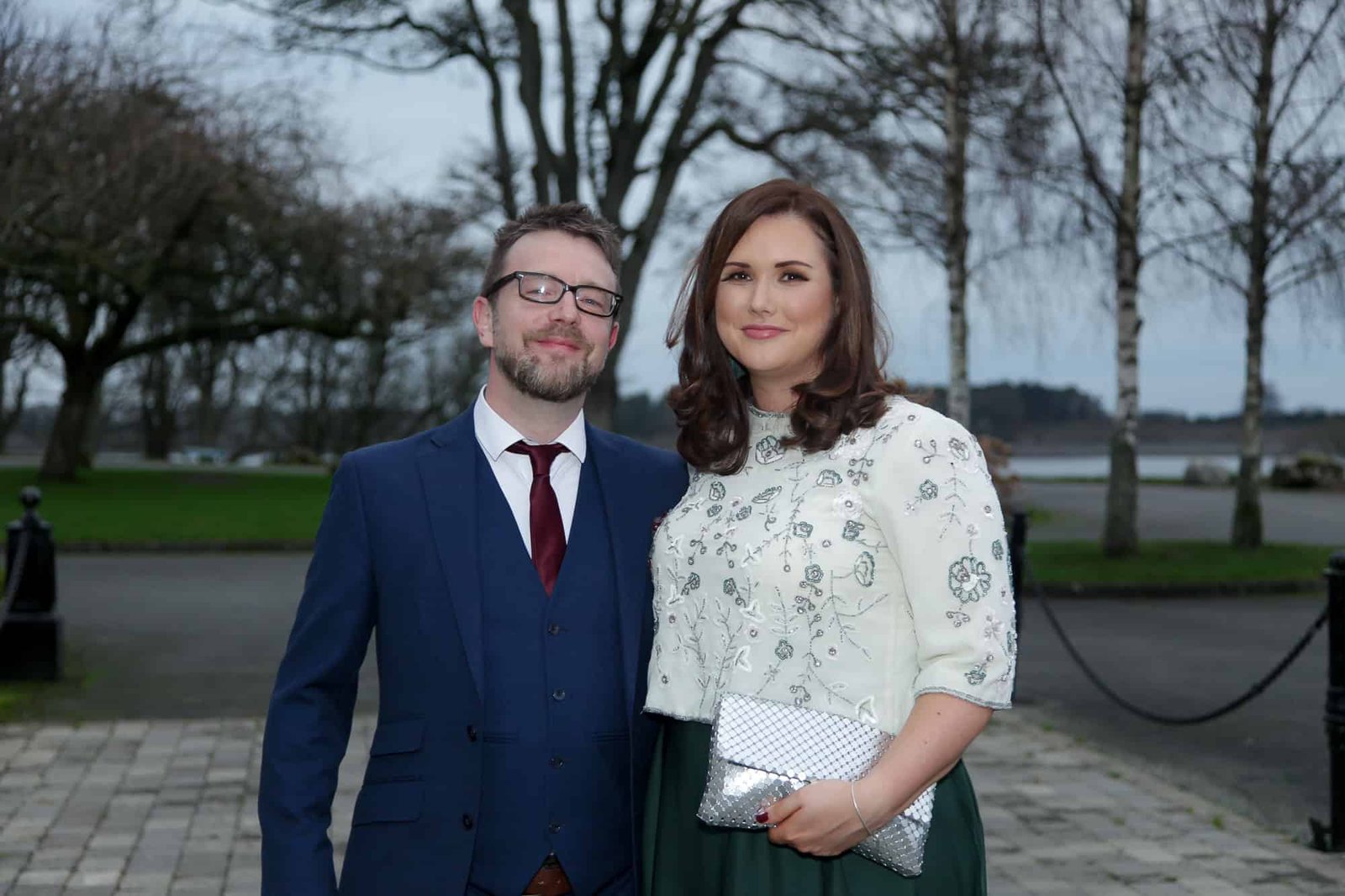 Elegant couple at outdoor event, standing by lake with trees in background.