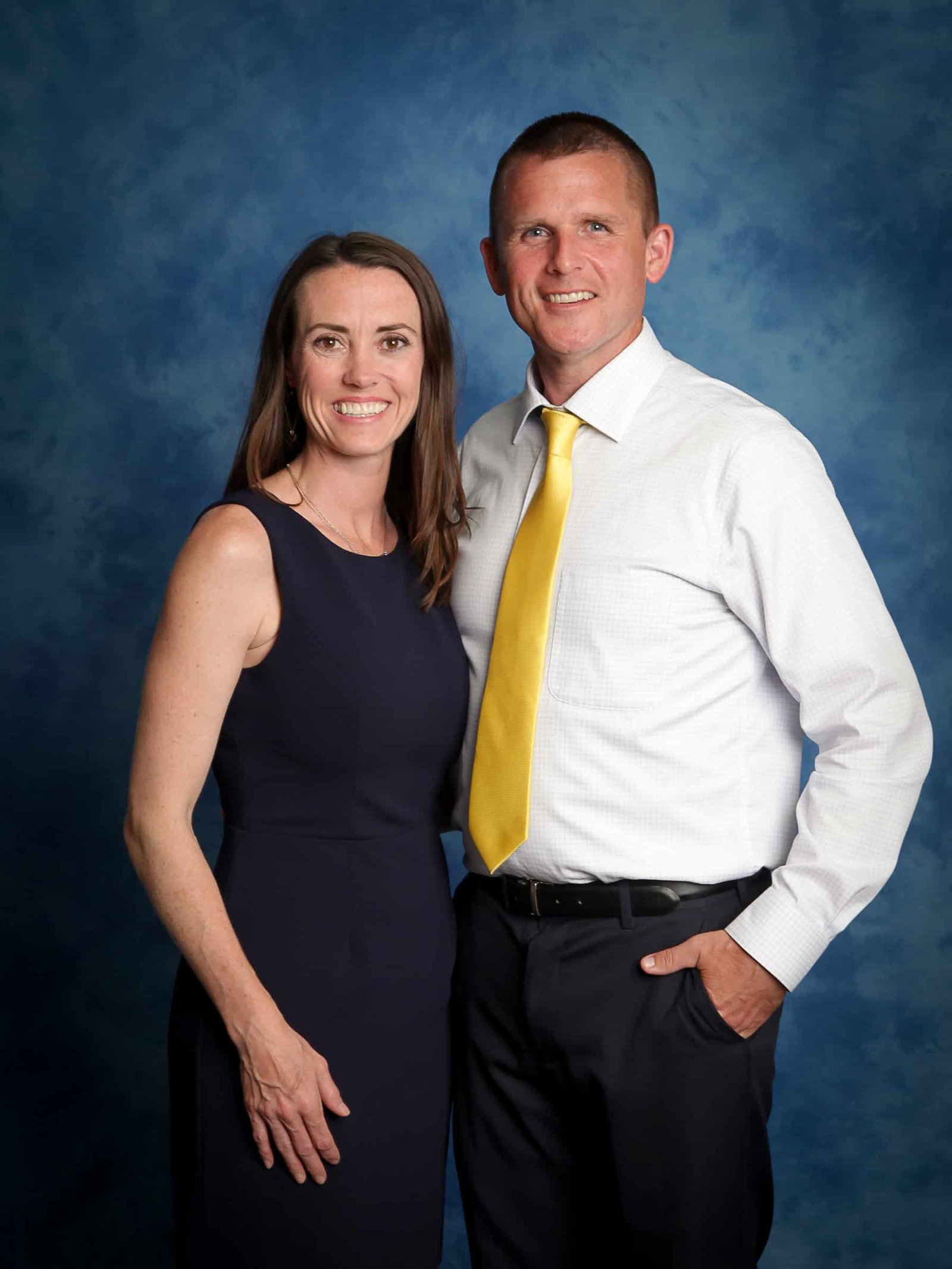 A woman in a navy blue dress and a man in a white shirt with a yellow tie stand together against a blue textured background. They are smiling and facing the camera.