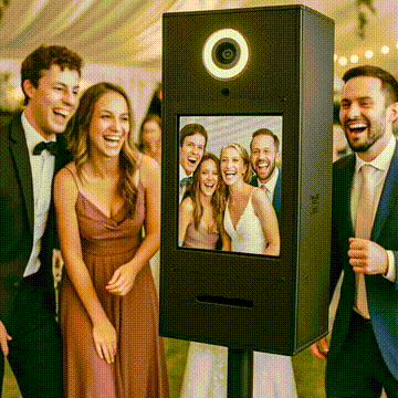 Four well-dressed people stand and smile next to a photo booth at an event. The photo booth screen displays their group photo, showing them laughing together in the same outfits.
