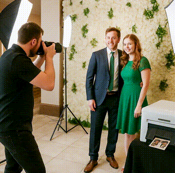 A photographer takes a picture of a man in a suit and a woman in a green dress standing in front of a flower wall backdrop, with photo equipment and a small printer visible nearby.