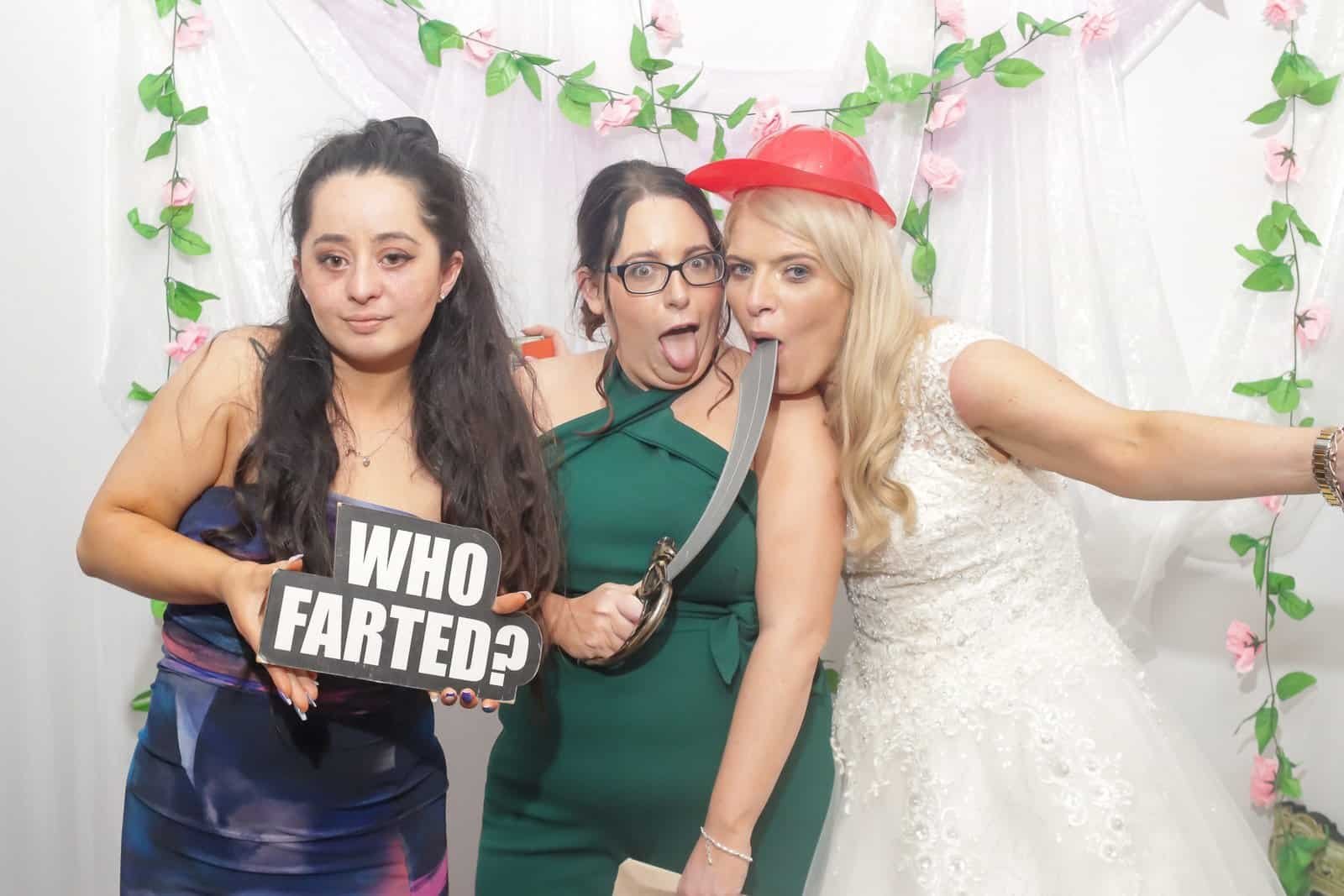 Three women at a photo booth; one holds a "WHO FARTED?" sign, another is wearing glasses and holding a prop, and the third, dressed as a bride, pretends to lick a fake sword. Pink and green floral backdrop.