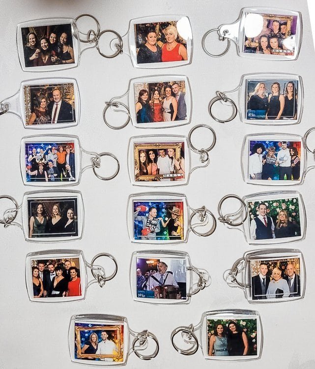 A collection of sixteen rectangular photo keyrings arranged on a white surface, each displaying groups of people posing and smiling at various social events.