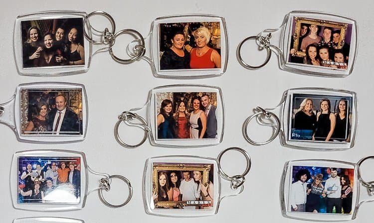 A display of nine rectangular keyrings, each containing a small photograph of groups of people posing together, arranged in three rows on a white surface.