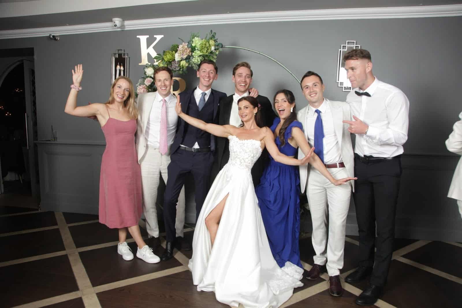 Eight people pose together indoors; a bride in a white dress stands front and centre, surrounded by friends in formal and semi-formal attire. The group smiles and gestures playfully in front of a grey wall with decorative elements.